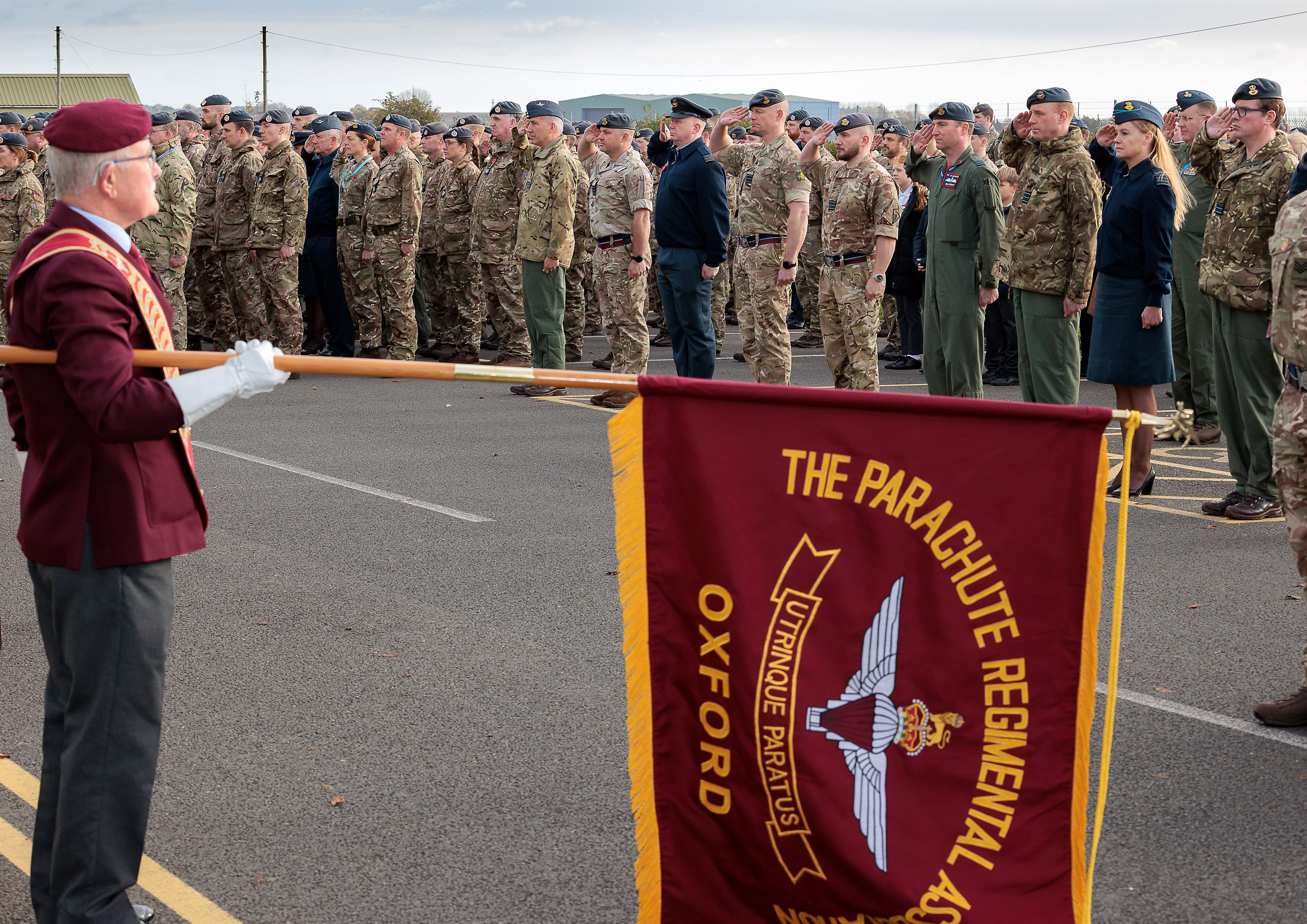 Photo: Foreground - Veteran holding a lowered Parachute Regimental Association of Oxford flag in front of gathered Whole Force members